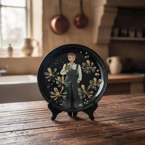 Può includere: Un piatto rotondo in ceramica nera con un'illustrazione centrale di una persona che indossa una salopette scura e una camicia bianca, mentre tiene in mano un piccolo bouquet. Il piatto è decorato con motivi floreali gialli e bianchi e poggia su un supporto nero.