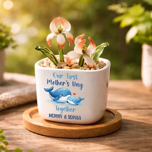 May include: A white ceramic planter with the text "Our First Mother's Day" and a whale illustration. The planter contains decorative flowers with white and orange petals. The planter sits on a wooden coaster.