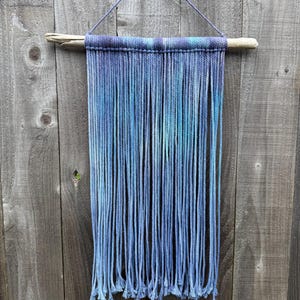 May include: A blue and purple macrame wall hanging. The top is a woven band of blue and purple yarn, attached to a natural wooden dowel. Long strands of blue yarn hang down, creating a textured, fringed effect. The wall hanging is suspended by a blue string.