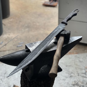 Può includere: Una spada nera con un manico di legno è appoggiata su un'incudine di metallo. Un martello di legno è anche sull'incudine.