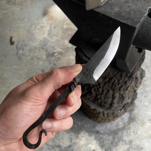 May include: A hand holds a small, black, forged knife with a curved handle and a sharp, silver blade. The knife is resting on a black anvil.