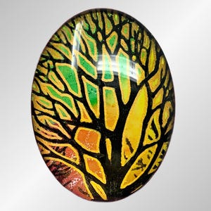 May include: An oval-shaped glass cabochon featuring a stylized tree design. The tree's branches and trunk are black, with vibrant yellow, green, and orange hues filling the spaces. The cabochon has a glossy finish and is set against a gradient gray background.