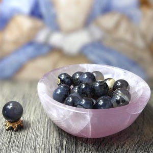 May include: A collection of dark blue, polished gemstone spheres in a light pink, oval-shaped bowl. One sphere rests on a small, gold-colored stand. The bowl and stand are on a wooden surface.