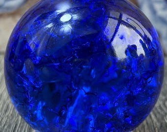 May include: A deep blue, spherical crystal ball with internal patterns, resting on a light-colored, circular stand. The sphere reflects light, creating a shimmering effect. The background features a painting of a person in a blue dress.