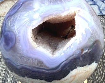 May include: A polished, spherical geode with a hollow center, showcasing bands of purple, white, and dark blue. The geode sits on a light-colored, intricately carved wooden stand, displaying a natural, earthy aesthetic.