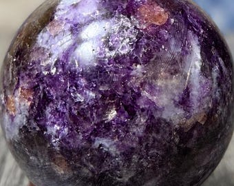 May include: A polished spherical crystal featuring deep purple and translucent white mineral inclusions throughout its surface. The sphere is mounted on a small, circular, dark brown wooden stand that provides a stable base for the stone.