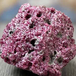 May include: A close-up of a rough, textured, pink mineral specimen. The rock has a porous appearance with several small holes and a slightly uneven surface. The color is a vibrant, deep pink with some areas of gray.