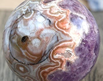 May include: A polished, spherical gemstone with a swirling pattern of white, orange, and brown against a purple background. The sphere is displayed on a small, light-colored wooden stand. The gemstone has a glossy finish.