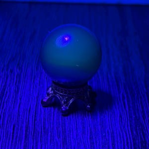 May include: A green crystal ball with a small, glowing red center sits atop an ornate, silver-colored stand. The ball is illuminated by a blue light, creating a mystical and ethereal effect. The stand has a detailed, antique design.