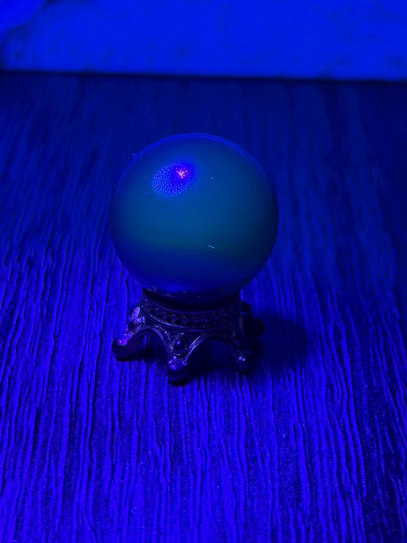 May include: A green crystal ball with a small, glowing red center sits atop an ornate, silver-colored stand. The ball is illuminated by a blue light, creating a mystical and ethereal effect. The stand has a detailed, antique design.