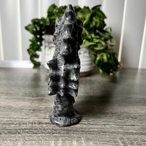 以下が含まれることがあります： テクスチャ加工された有機的なデザインの、グレーと黒の石の彫刻。彫刻は、丸いベースと尖った上部を備えた、高く垂直な形状をしています。背景には、白い鉢植えの緑の植物があります。