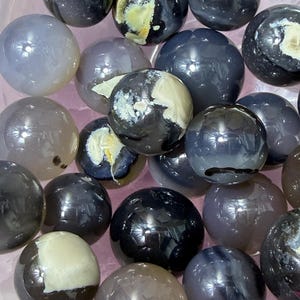 May include: A collection of polished gemstone spheres in various shades of gray, black, and cream. The spheres have a glossy, reflective surface and are arranged closely together. Some spheres have unique patterns and color variations.