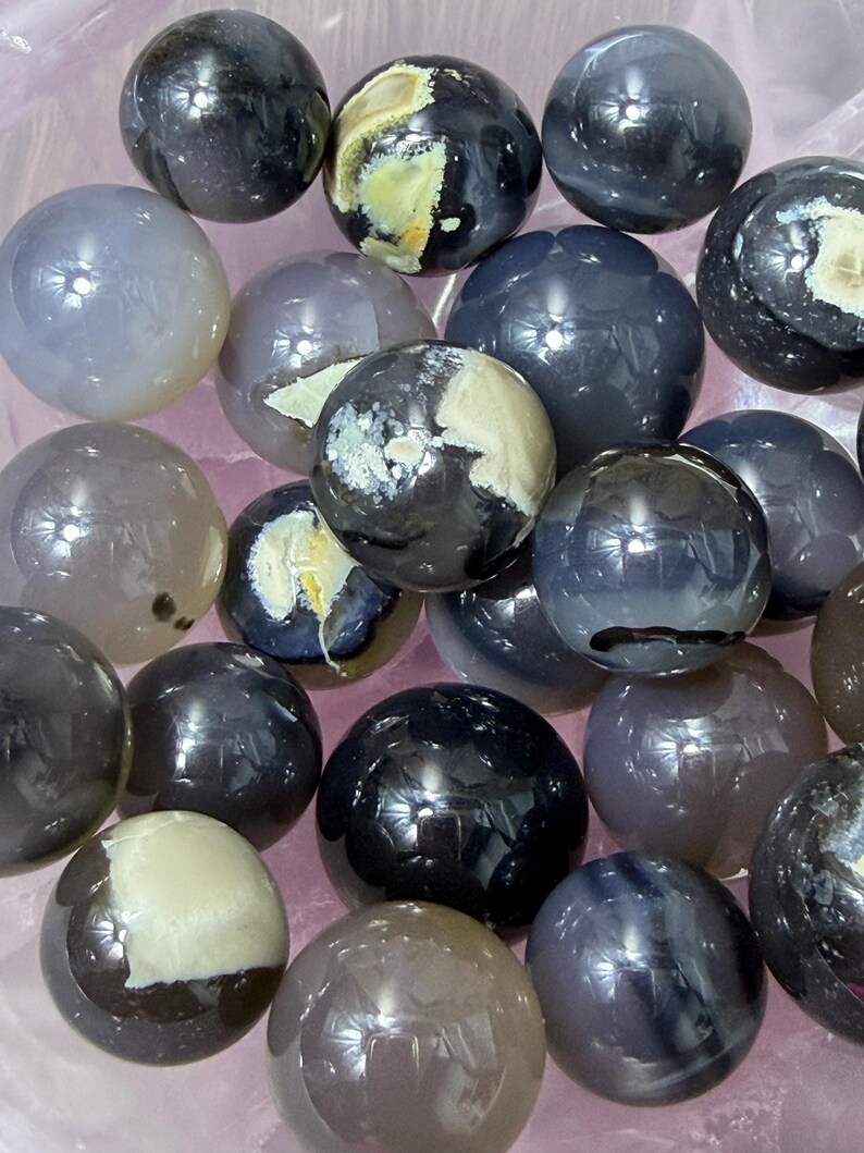 May include: A collection of polished gemstone spheres in various shades of gray, black, and cream. The spheres have a glossy, reflective surface and are arranged closely together. Some spheres have unique patterns and color variations.