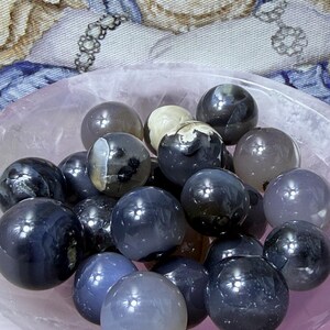 May include: A collection of polished gemstone spheres in shades of gray, blue, and white, filling a light pink bowl. The spheres vary in size and have a glossy finish, reflecting light. The background features a patterned fabric.