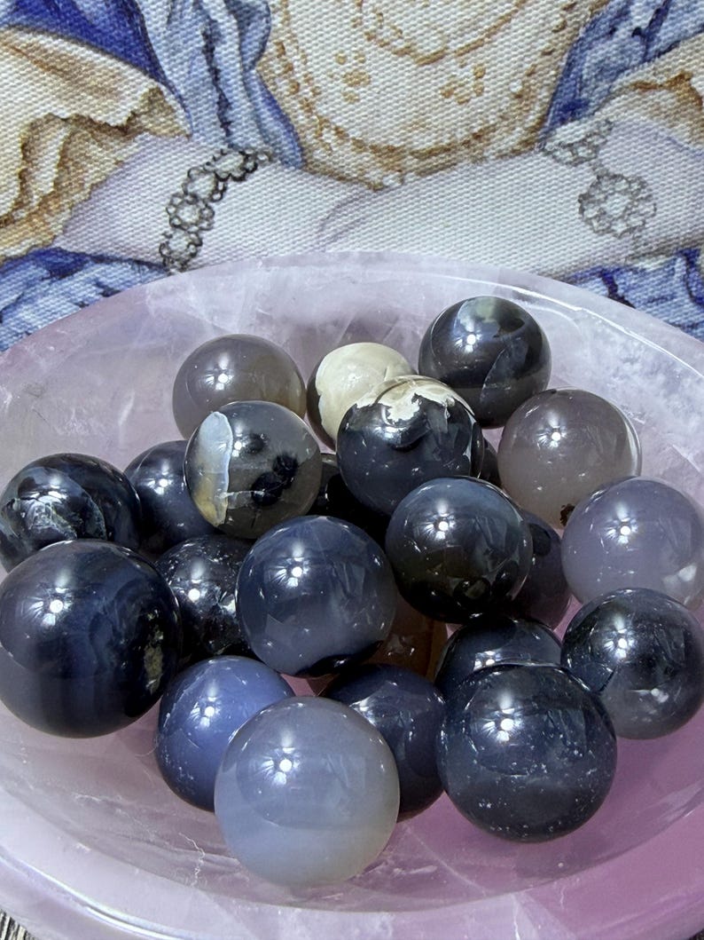 May include: A collection of polished gemstone spheres in shades of gray, blue, and white, filling a light pink bowl. The spheres vary in size and have a glossy finish, reflecting light. The background features a patterned fabric.