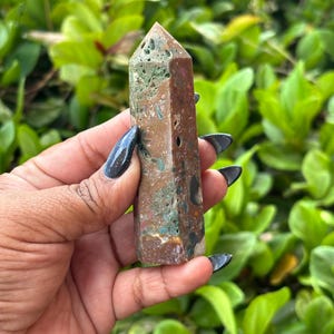 May include: A natural stone obelisk with a pointed top. The stone features a mix of brown, green, and tan colors. The obelisk is held against a backdrop of green foliage.