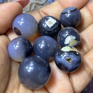 May include: A collection of polished, spherical gemstones in shades of gray and blue. Some spheres have unique patterns and internal structures, including white and brown inclusions. The gemstones are held in a hand, showcasing their smooth, reflective surfaces.