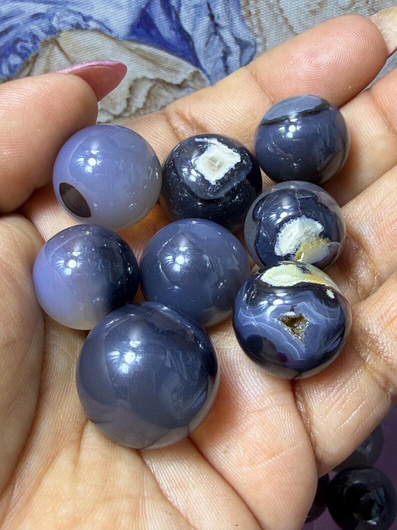 May include: A collection of polished, spherical gemstones in shades of gray and blue. Some spheres have unique patterns and internal structures, including white and brown inclusions. The gemstones are held in a hand, showcasing their smooth, reflective surfaces.