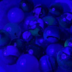 May include: A collection of colorful marbles in a white container, illuminated by a blue light. The marbles feature a variety of colors, including green, white, and black, with swirling patterns and solid hues. The image is taken from a close-up perspective.