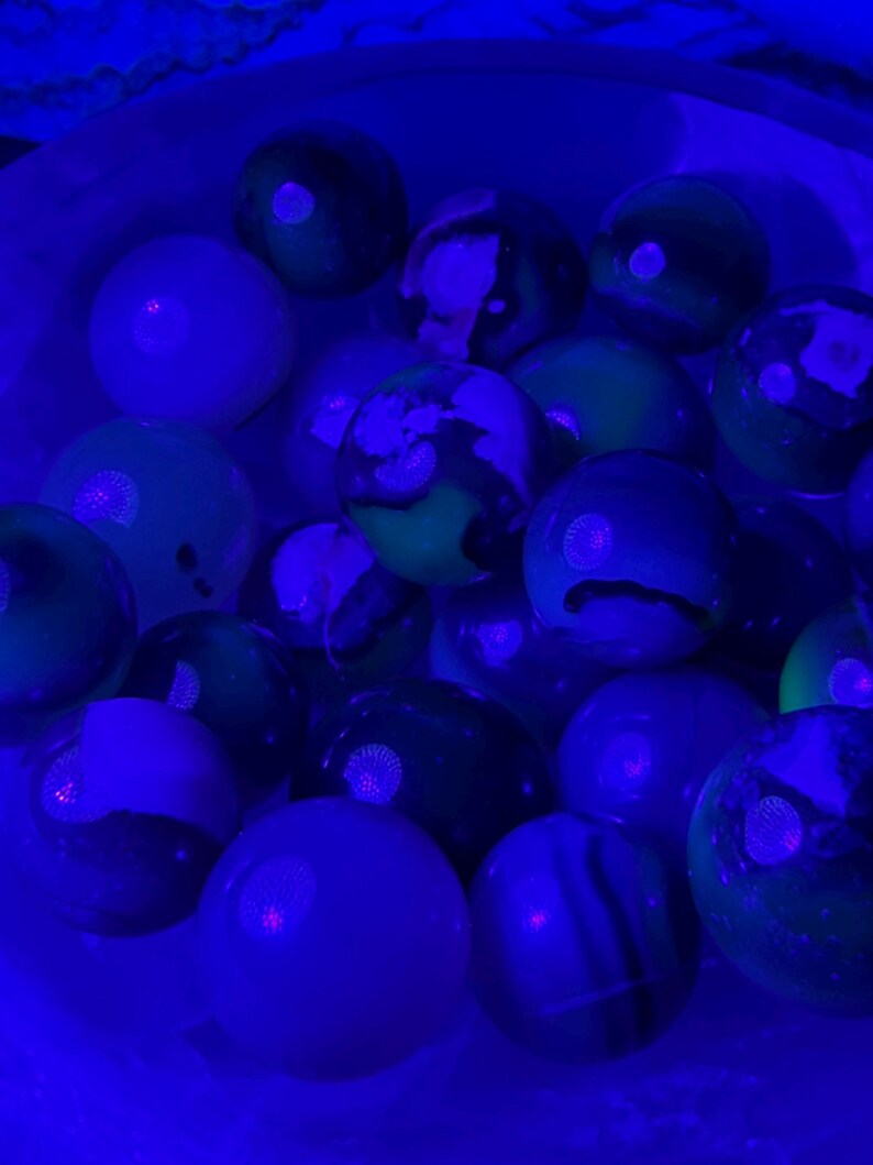 May include: A collection of colorful marbles in a white container, illuminated by a blue light. The marbles feature a variety of colors, including green, white, and black, with swirling patterns and solid hues. The image is taken from a close-up perspective.