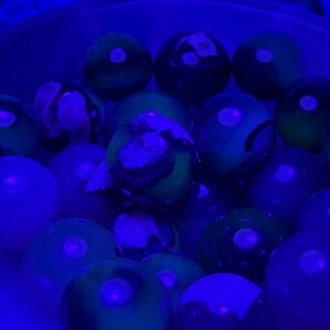 May include: A collection of glowing marbles in shades of green, blue, and purple. The marbles have a glossy finish and are illuminated by a blue light, creating a vibrant and otherworldly effect. The marbles are of varying sizes and have unique patterns.