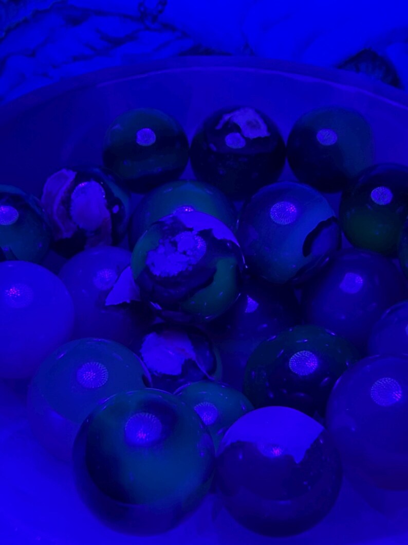 May include: A collection of glowing marbles in shades of green, blue, and purple. The marbles have a glossy finish and are illuminated by a blue light, creating a vibrant and otherworldly effect. The marbles are of varying sizes and have unique patterns.