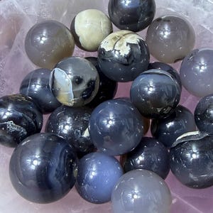 May include: A collection of polished gemstone spheres in various shades of gray, black, and blue, resting in a pink bowl. Some spheres have unique patterns and textures. The spheres vary in size and reflect light, creating a visually appealing display.