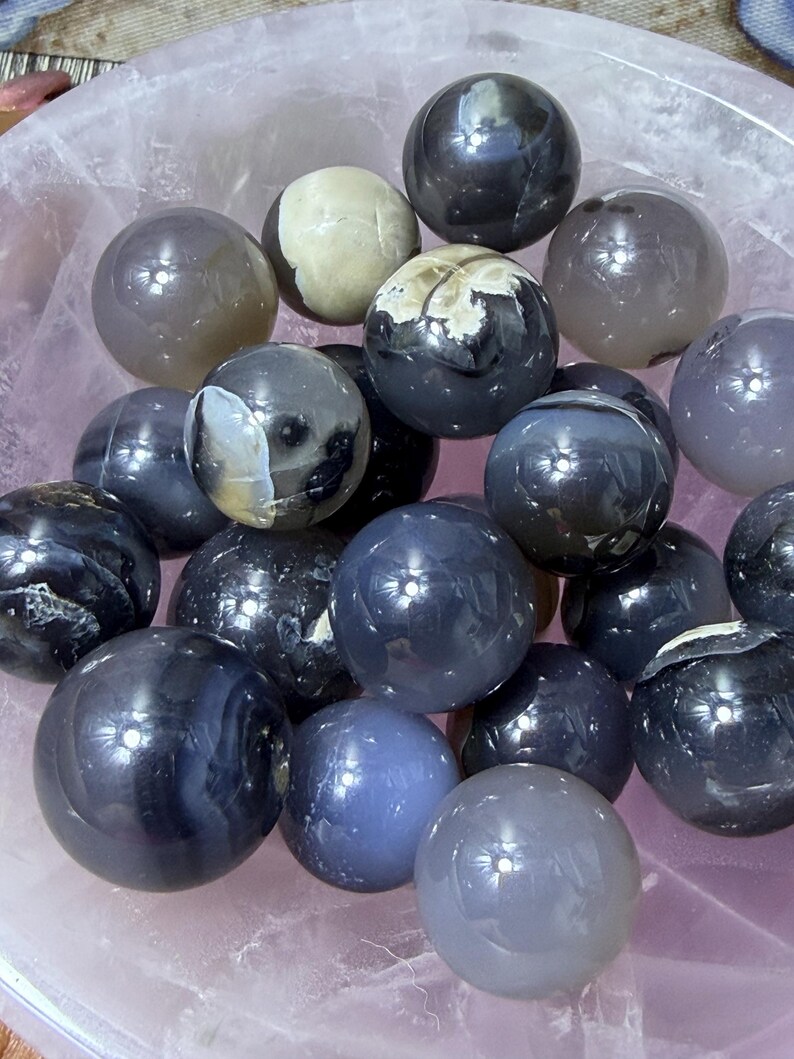 May include: A collection of polished gemstone spheres in various shades of gray, black, and blue, resting in a pink bowl. Some spheres have unique patterns and textures. The spheres vary in size and reflect light, creating a visually appealing display.