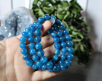 May include: A hand holds three strands of bright blue gemstone bead bracelets. The beads are round and have a polished, reflective surface. The background includes a light blue crystal and green foliage.