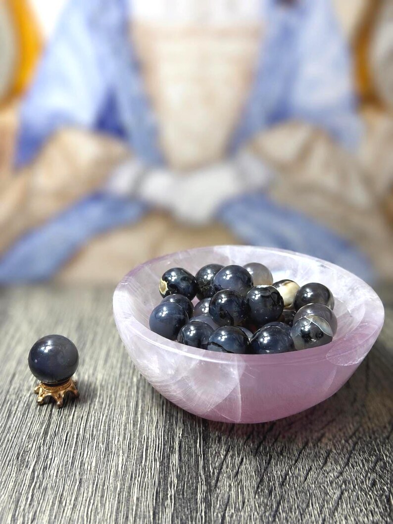May include: A collection of dark blue, polished gemstone spheres in a light pink, oval-shaped bowl. One sphere rests on a small, gold-colored stand. The bowl and stand are on a wooden surface.