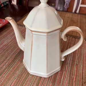 May include: A white ceramic teapot with a brown trim and a lid. The teapot has a rounded body and a spout on the side. The teapot is sitting on a brown and green placemat.