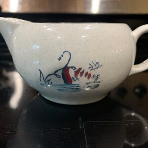 May include: White ceramic creamer with a blue and red swan design. The creamer has a small handle and a rounded shape.