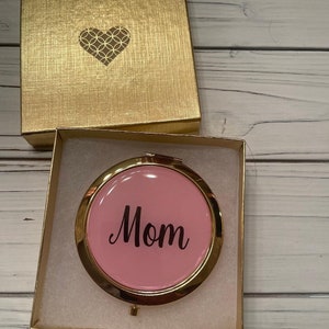 May include: A gold-colored gift box with a heart design on the lid, open to reveal a pink compact mirror. The mirror has a gold rim and the word "Mom" in black script.