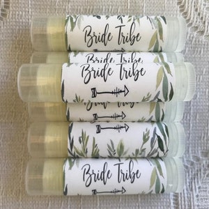 May include: A stack of clear lip balm tubes with white labels. The labels have a green watercolour floral design and the text "Bride Tribe" in black.