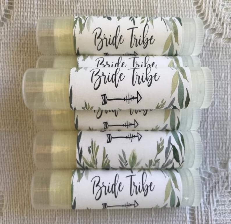 May include: A stack of clear lip balm tubes with white labels. The labels have a green watercolour floral design and the text "Bride Tribe" in black.