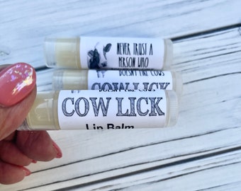 May include: Three cylindrical lip balm tubes with white labels. The labels feature a black and white cow illustration and the text NEVER TRUST A PERSON WHO DOESN'T LIKE COWS and COW LICK Lip Balm in a bold, stylized black font on a clear plastic tube.