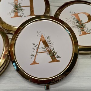 May include: Four gold compact mirrors with white lids and gold lettering. Each mirror features a different letter of the alphabet, A, H, and P, with gold leaf and green foliage designs.
