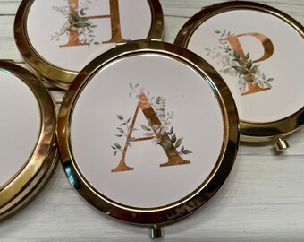 May include: Four gold compact mirrors with white lids and gold lettering. Each mirror features a different letter of the alphabet, A, H, and P, with gold leaf and green foliage designs.