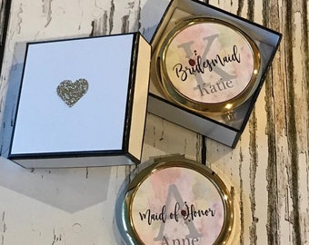 May include: Two compact mirrors with gold rims and watercolor designs. One mirror reads "Bridesmaid Katie" and the other reads "Maid of Honor Anne." A white box with a black trim and a glitter heart is also in the image.