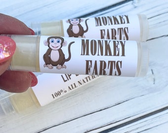 May include: Clear lip balm tubes with white labels featuring a cartoon monkey and the words "MONKEY FARTS." The text "LIP BALM" and "100% ALL NATURAL" are also visible. The lip balm is held by a person.