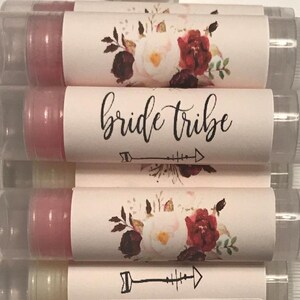 May include: A stack of clear tubes containing pink lip balm. Each tube has a white label with a floral design and the text "bride tribe" with an arrow underneath.