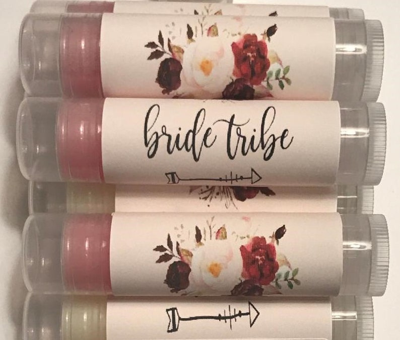 May include: A stack of clear tubes containing pink lip balm. Each tube has a white label with a floral design and the text "bride tribe" with an arrow underneath.