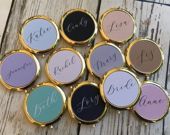 May include: A collection of personalized compact mirrors with gold-colored rims. Each mirror features a different pastel-colored background and a name written in elegant script. The names include "Katie", "Cindy", "Lisa", "Jennifer", "Rachel", "Mary", "Liz", "Beth", "Lory", "Bride", and "Anne".
