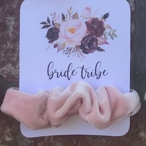 May include: A white card with a floral design and the text "bride tribe" printed on it. A pink velvet scrunchie is placed on top of the card.