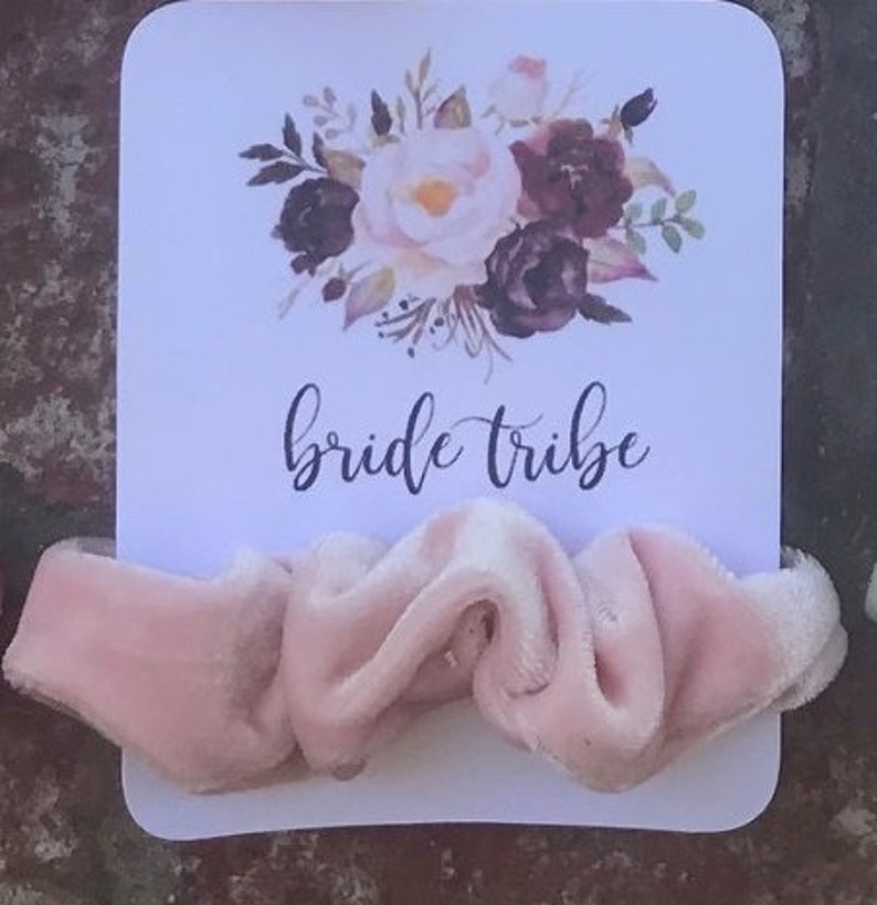 May include: A white card with a floral design and the text "bride tribe" printed on it. A pink velvet scrunchie is placed on top of the card.
