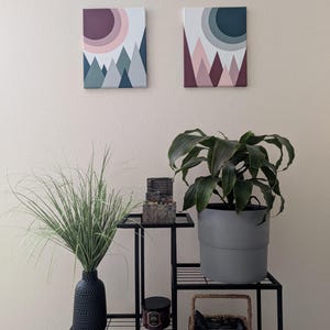 May include: Two framed canvas art pieces depicting stylized mountain landscapes with a sun. The color palettes include shades of pink, purple, and blue. A black metal plant stand holds a variety of decorative items and plants.