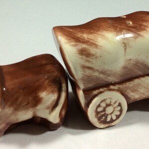 May include: A brown and white ceramic salt and pepper shaker set. The salt shaker is shaped like an ox pulling a covered wagon. The wagon has two wheels and is painted brown and white.