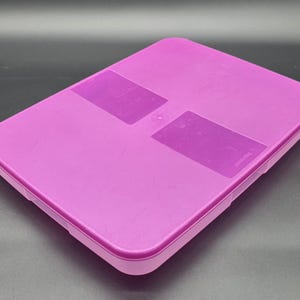 Puede incluir: Un recipiente rectangular de color magenta con una tapa a juego. La tapa presenta dos parches rectangulares de color magenta más oscuro. El recipiente está hecho de un material liso y sólido y está diseñado para el almacenamiento.