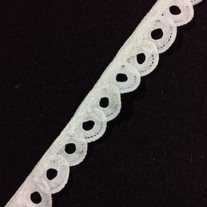 May include: White lace trim with a scalloped edge and circular cutouts. The delicate trim is angled across a black background, showcasing its intricate design. Ideal for sewing, crafting, or adding a decorative touch to garments or home decor projects.
