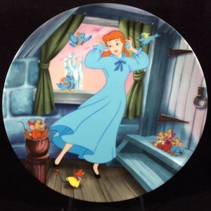 May include: A blue and white porcelain plate featuring a depiction of Cinderella in a blue nightgown, surrounded by mice and birds. The scene takes place in a bedroom with a window overlooking a castle.